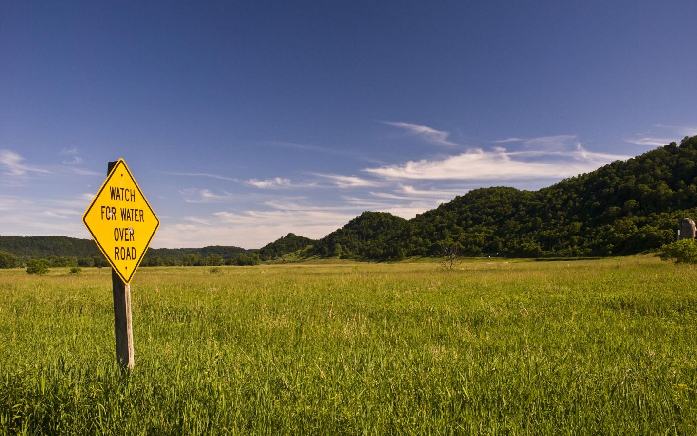 "Watch For Water Over Road" sign in a grassy field